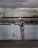 A person stands on a concrete walkway by the water, holding a fishing line near a fishing rod. They are wearing a cap, a brown and white striped shirt, and camouflage shorts. The background features a large, industrial building and a crane under a cloudy sky.