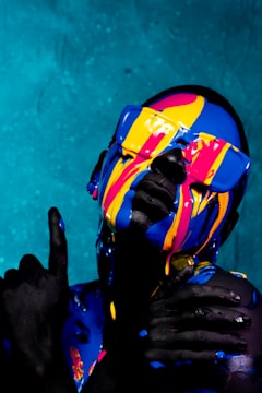A person is covered in vibrant, dripping paint, with bold colors of blue, yellow, and pink. The background is a textured teal surface. The paint streams down the face and sunglasses, creating a striking contrast against the skin. The person’s pose conveys confidence and creativity.