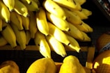 A large pile of ripe yellow bananas is arranged on a wooden shelf with sunlight casting shadows over the fruit. The bananas are closely packed together, and there are hints of other fruits or objects in the background.