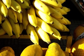 A large pile of ripe yellow bananas is arranged on a wooden shelf with sunlight casting shadows over the fruit. The bananas are closely packed together, and there are hints of other fruits or objects in the background.