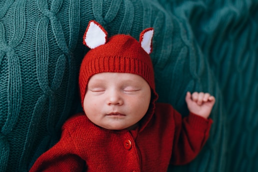 A sleeping baby is dressed in a red knitted outfit with fox-like ears on the hat, lying on a textured green knitted surface.