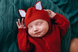 A newborn baby is peacefully sleeping, dressed in a red knitted outfit with tiny ear-like decorations on the hat. The baby is lying on a textured green blanket, creating a cozy and warm atmosphere.
