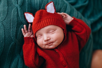 A newborn baby is peacefully sleeping, dressed in a red knitted outfit with tiny ear-like decorations on the hat. The baby is lying on a textured green blanket, creating a cozy and warm atmosphere.