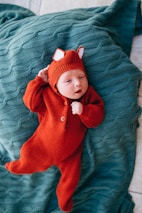 A baby dressed in an orange-red knitted outfit with fox ears lies on a textured teal-green blanket.