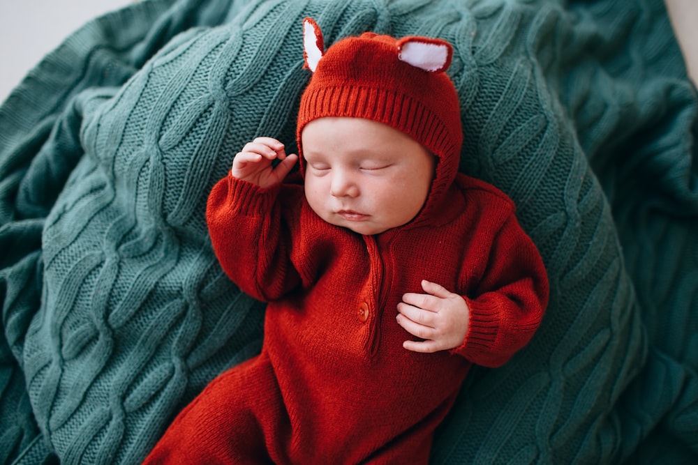baby in red knit cap and red sweater lying on green textile