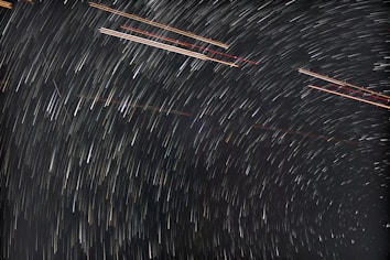 Long exposure photograph of the night sky showcasing star trails forming circular patterns. Lines of varying brightness and color arc across the image, indicating the apparent motion of stars over time.
