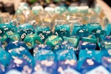 A collection of colorful, multi-sided gaming dice with numbers, predominantly in shades of blue and green, arranged closely and reflecting light.