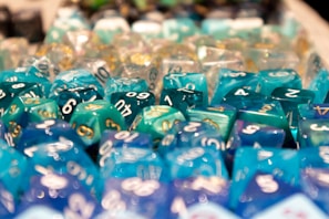 A collection of colorful, multi-sided gaming dice with numbers, predominantly in shades of blue and green, arranged closely and reflecting light.