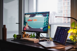 A workspace featuring a desk with a desktop monitor and a laptop. The monitor displays an illustrated scene with a cartoon animal in a boat on a lake surrounded by trees. Several items are on the desk, including a brown bottle, a small Android figurine, and a plush toy. Potted plants and modern building windows are visible in the background.