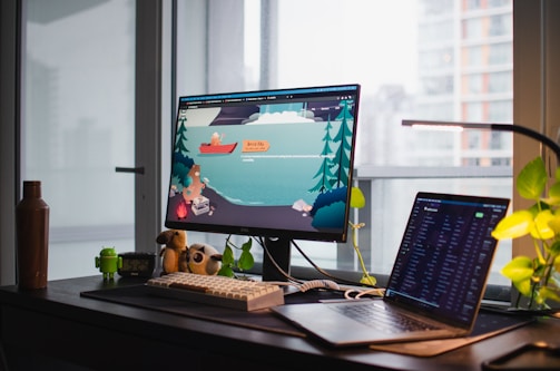 A workspace featuring a desk with a desktop monitor and a laptop. The monitor displays an illustrated scene with a cartoon animal in a boat on a lake surrounded by trees. Several items are on the desk, including a brown bottle, a small Android figurine, and a plush toy. Potted plants and modern building windows are visible in the background.