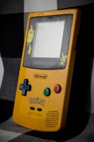 A yellow Game Boy Color featuring Pikachu graphics on the faceplate rests against a checkered fabric background. Buttons include a blue directional pad and green and red action buttons, with a worn exterior suggesting age and use.