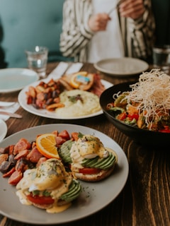 A table set with an inviting brunch assortment, featuring eggs Benedict with avocado and hollandaise sauce on a gray plate accompanied by roasted potatoes and a slice of orange. A colorful bowl of stir-fried vegetables garnished with crispy noodles is also present. A person in a striped shirt is visible in the background, focusing on their meal preparation.