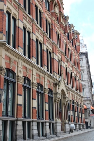 A historic building with intricate architectural details, featuring orange bricks and beige stone elements. The structure includes tall, narrow windows with dark frames and decorative elements along the ledges and columns.