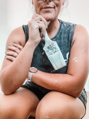 A person with crossed arms is holding a paintbrush covered in white paint. The individual has short hair tied back and is wearing a sleeveless dark shirt adorned with paint splatters. A watch is visible on one wrist, and there are rings on both hands.
