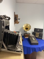 A room displays various vintage items including a large format camera, an old telephone mounted on the wall, a gramophone with a brass horn, a Victor adding machine, and a vintage radio. The objects are placed on a table covered with a blue cloth, with the camera positioned in the foreground.