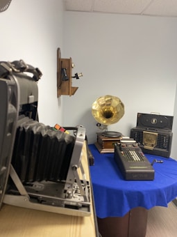 A room displays various vintage items including a large format camera, an old telephone mounted on the wall, a gramophone with a brass horn, a Victor adding machine, and a vintage radio. The objects are placed on a table covered with a blue cloth, with the camera positioned in the foreground.