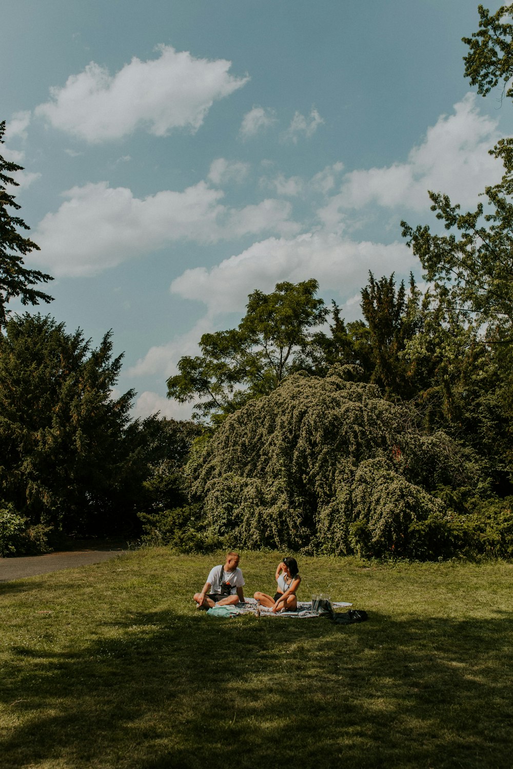 2 women sitting on green grass field near green trees under white clouds and blue sky