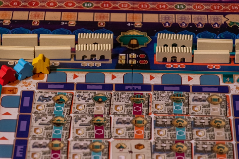 A colorful board game layout with intricate patterns and game pieces. The board features rows and columns with different symbols and tracks, including small structures resembling buildings and pawn-like figures in blue, red, and yellow.