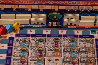A colorful board game layout with intricate patterns and game pieces. The board features rows and columns with different symbols and tracks, including small structures resembling buildings and pawn-like figures in blue, red, and yellow.