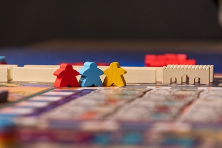 Colorful board game pieces are arranged on a game board. Three key pieces, shaped like small figures, are positioned prominently in red, blue, and yellow. In the background, a white structure reminiscent of a wall or castle adds an element of architecture to the scene. The game board is partially visible, showing a mosaic of colors and patterns.