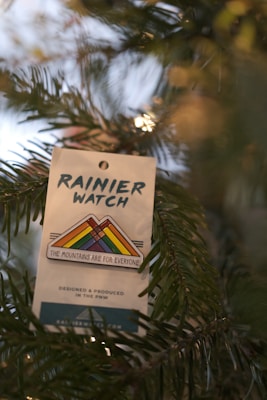 A label or tag is attached to a branch of a pine tree. The tag displays the text 'RAINIER WATCH' followed by a multicolored mountain logo with the message 'THE MOUNTAINS ARE FOR EVERYONE'. The environment suggests a natural setting, possibly outdoors or a holiday-themed decoration.