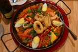 A large pan filled with a vibrant paella, featuring yellow rice, shrimp, fried calamari rings, hard-boiled egg wedges, peas, red bell peppers, and corn. A spoon rests on the right side of the pan, and there's a plate and a bottle partially visible in the background.
