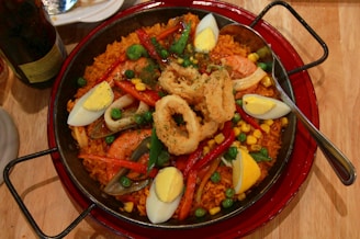 A large pan filled with a vibrant paella, featuring yellow rice, shrimp, fried calamari rings, hard-boiled egg wedges, peas, red bell peppers, and corn. A spoon rests on the right side of the pan, and there's a plate and a bottle partially visible in the background.