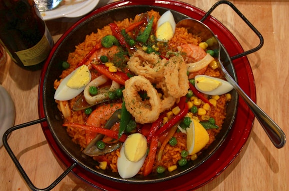 A large pan filled with a vibrant paella, featuring yellow rice, shrimp, fried calamari rings, hard-boiled egg wedges, peas, red bell peppers, and corn. A spoon rests on the right side of the pan, and there's a plate and a bottle partially visible in the background.
