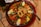 A large pan filled with a vibrant paella, featuring yellow rice, shrimp, fried calamari rings, hard-boiled egg wedges, peas, red bell peppers, and corn. A spoon rests on the right side of the pan, and there's a plate and a bottle partially visible in the background.