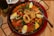 A large pan filled with a vibrant paella, featuring yellow rice, shrimp, fried calamari rings, hard-boiled egg wedges, peas, red bell peppers, and corn. A spoon rests on the right side of the pan, and there's a plate and a bottle partially visible in the background.