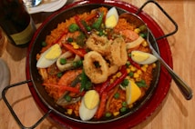 A large pan filled with a vibrant paella, featuring yellow rice, shrimp, fried calamari rings, hard-boiled egg wedges, peas, red bell peppers, and corn. A spoon rests on the right side of the pan, and there's a plate and a bottle partially visible in the background.