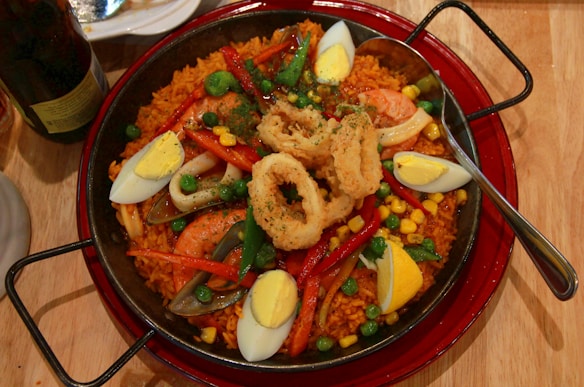 A large pan filled with a vibrant paella, featuring yellow rice, shrimp, fried calamari rings, hard-boiled egg wedges, peas, red bell peppers, and corn. A spoon rests on the right side of the pan, and there's a plate and a bottle partially visible in the background.