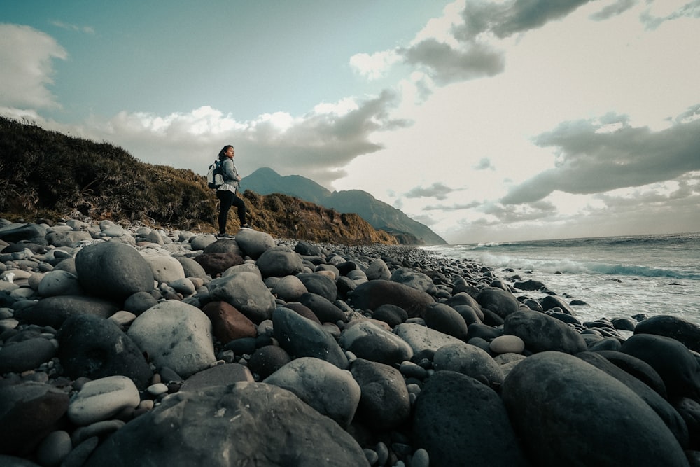 person in black jacket and blue denim jeans standing on rocky shore during daytime