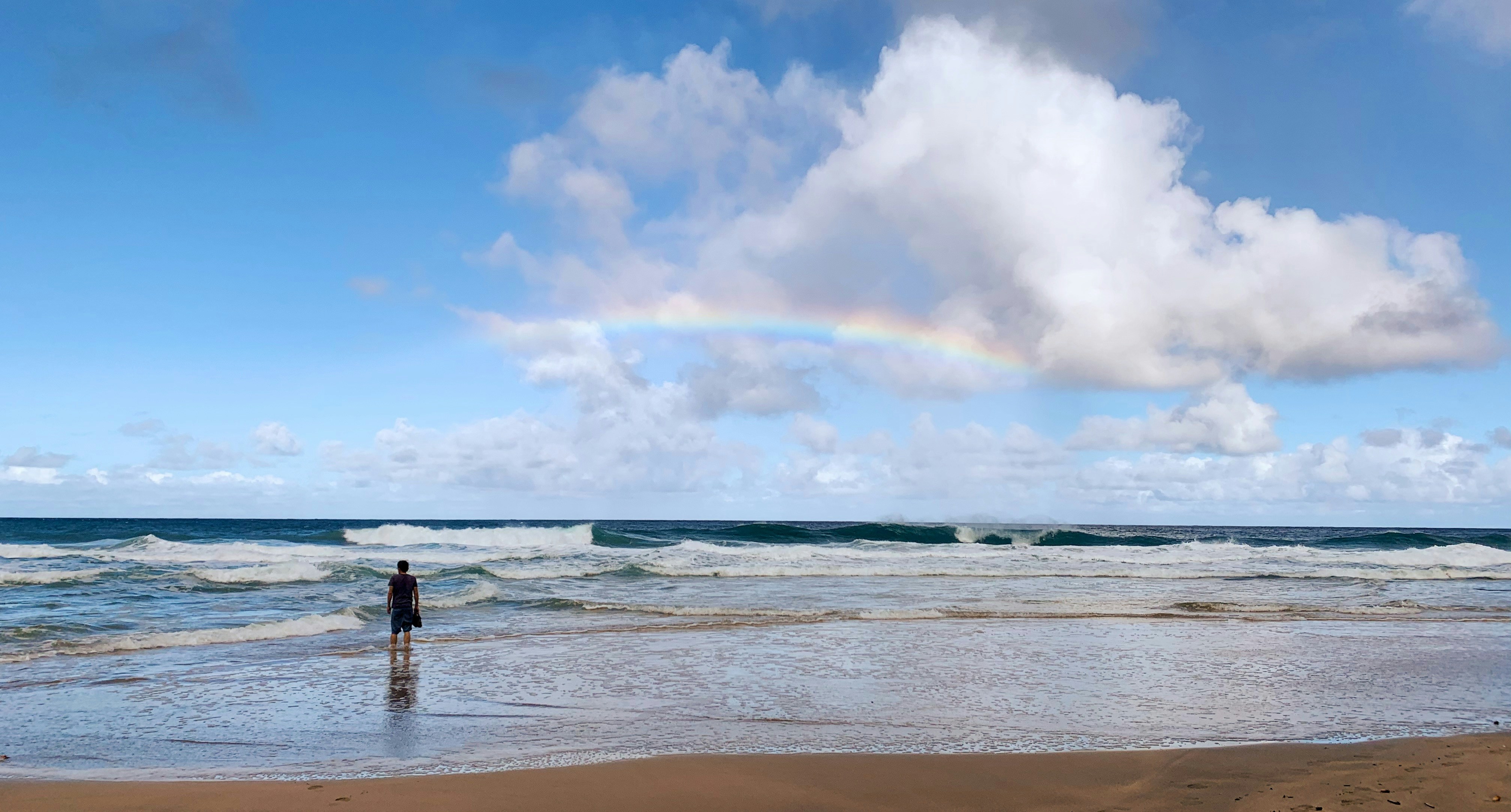 Man standing on beach under rainbow.