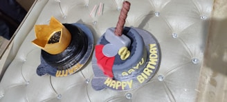 Two themed birthday cakes are placed on a cushioned surface. One cake is designed like a black hat with a golden crown on top, while the other resembles a hammer with a brown handle, a red base, and gray head, referencing a popular comic theme. 'WIMAL' is written on the first cake and 'AAVU HAPPY BIRTHDAY' on the second.
