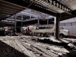A construction site with several workers and machinery in use. The area appears to be partially covered by a steel structure with beams overhead. A large cement mixer truck and another industrial vehicle are present. Workers in helmets are scattered around the muddy construction site.