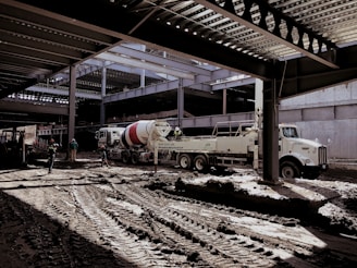 A construction site with several workers and machinery in use. The area appears to be partially covered by a steel structure with beams overhead. A large cement mixer truck and another industrial vehicle are present. Workers in helmets are scattered around the muddy construction site.