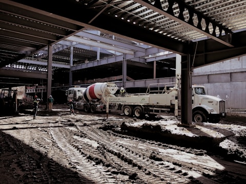 A construction site with several workers and machinery in use. The area appears to be partially covered by a steel structure with beams overhead. A large cement mixer truck and another industrial vehicle are present. Workers in helmets are scattered around the muddy construction site.