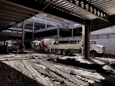 A construction site with several workers and machinery in use. The area appears to be partially covered by a steel structure with beams overhead. A large cement mixer truck and another industrial vehicle are present. Workers in helmets are scattered around the muddy construction site.
