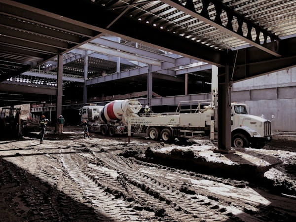 A construction site with several workers and machinery in use. The area appears to be partially covered by a steel structure with beams overhead. A large cement mixer truck and another industrial vehicle are present. Workers in helmets are scattered around the muddy construction site.