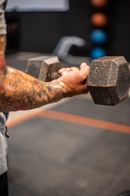 A person with tattooed arms is holding a large dumbbell, suggesting strength training. The focus is on their forearms and the weight, creating a sense of concentration and effort. In the blurred background, there are colorful medicine balls on a rack.