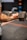A person with tattooed arms is holding a large dumbbell, suggesting strength training. The focus is on their forearms and the weight, creating a sense of concentration and effort. In the blurred background, there are colorful medicine balls on a rack.
