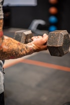 A person with tattooed arms is holding a large dumbbell, suggesting strength training. The focus is on their forearms and the weight, creating a sense of concentration and effort. In the blurred background, there are colorful medicine balls on a rack.