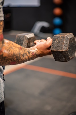 A person with tattooed arms is holding a large dumbbell, suggesting strength training. The focus is on their forearms and the weight, creating a sense of concentration and effort. In the blurred background, there are colorful medicine balls on a rack.