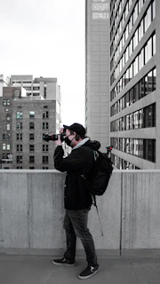 A person with a backpack is taking a photograph using a camera on an urban rooftop. The scene includes tall buildings with numerous windows lining the background, and the individual is dressed in casual clothing with a cap.