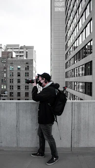 A person with a backpack is taking a photograph using a camera on an urban rooftop. The scene includes tall buildings with numerous windows lining the background, and the individual is dressed in casual clothing with a cap.