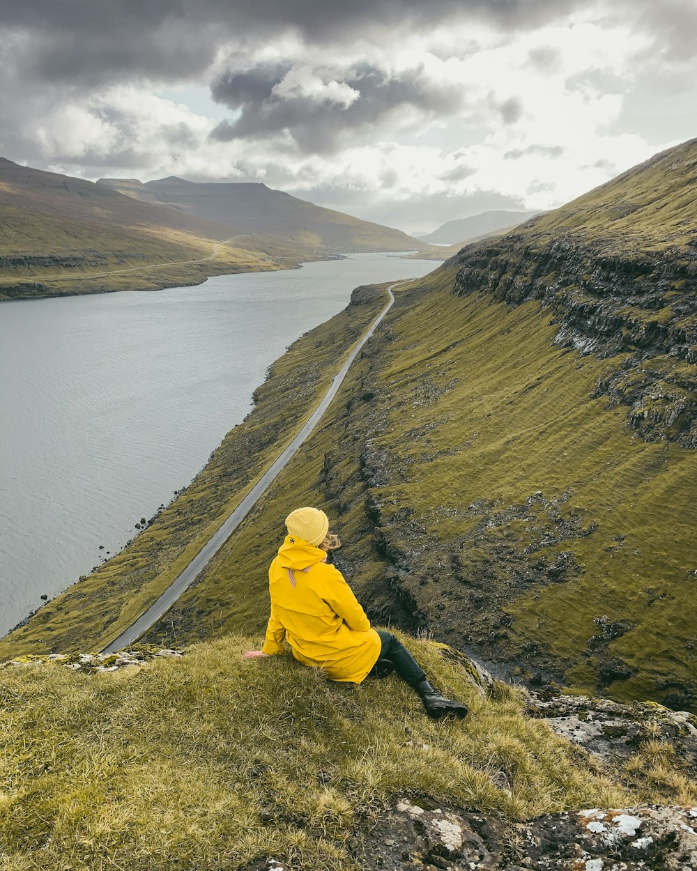 person in yellow hoodie sitting on rock near body of water during daytime