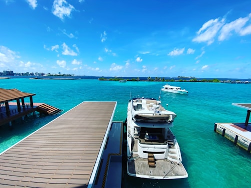 A luxurious boat is docked at a wooden pier on clear turquoise water under a bright blue sky with scattered clouds. Another smaller boat is seen floating nearby on the open sea. In the background, there is a cityscape with tall buildings and lush green islands.