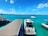 A luxurious boat is docked at a wooden pier on clear turquoise water under a bright blue sky with scattered clouds. Another smaller boat is seen floating nearby on the open sea. In the background, there is a cityscape with tall buildings and lush green islands.