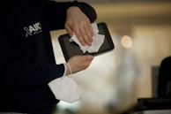 A person is cleaning a tablet with a white cloth while holding a white face mask in the other hand. The individual is wearing a dark blue shirt. The background is blurred, suggesting an indoor setting.